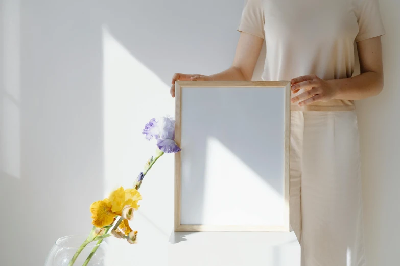 a woman holding a picture frame in front of a vase of flowers, a poster, pexels contest winner, visual art, natural window lighting, on a white table, backlight body, on a wooden tray