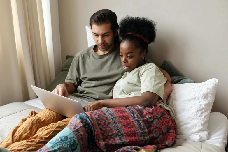 a man and woman sitting on a bed looking at a laptop, by Carey Morris, pexels, renaissance, light skinned african young girl, on a couch, multicoloured, cuddling