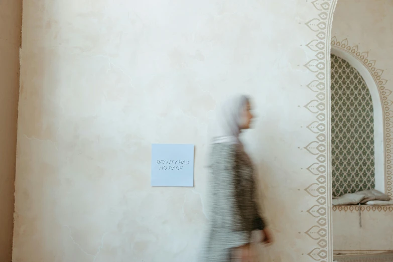 a woman walking past a wall with a sign on it, by Maryam Hashemi, trending on unsplash, visual art, he is in a mosque, silver and muted colors, lines and movement, background image