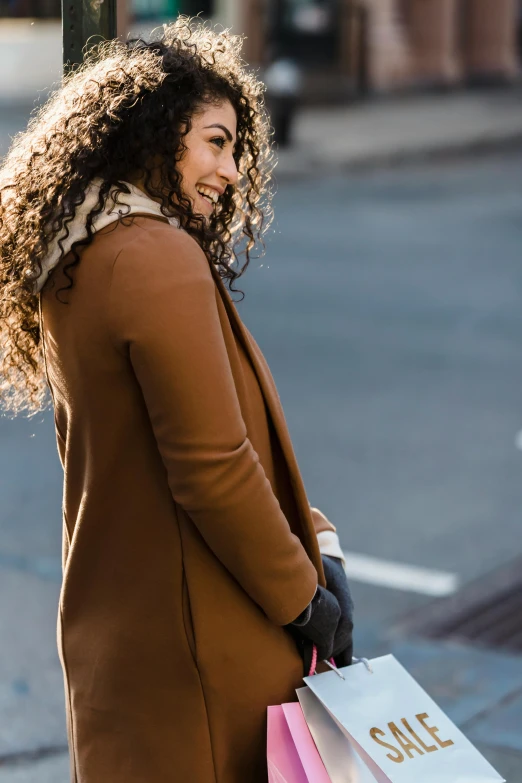 a woman walking down the street carrying shopping bags, a portrait, inspired by Anita Malfatti, trending on pexels, long dark curly hair, light brown coat, woman's profile, smiling