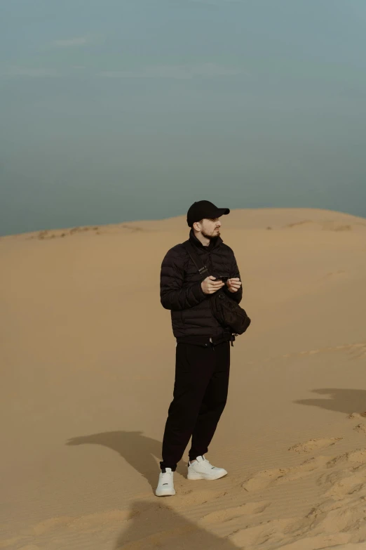 a man standing on top of a sand dune, an album cover, inspired by Amir Zand, trending on pexels, realism, mac miller, thoughtful pose, low quality, a man wearing a black jacket