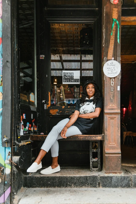 a woman sitting on a bench in front of a store, a portrait, by Bernie D’Andrea, pexels contest winner, black arts movement, graphic tees, sitting at a bar, wearing white sneakers, promotional image