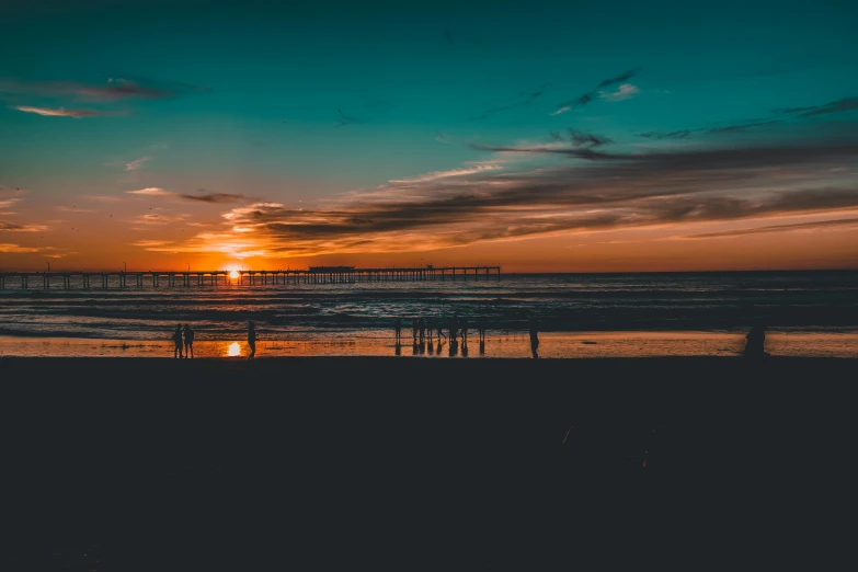 a group of people standing on top of a beach next to the ocean, pexels contest winner, minimalism, large twin sunset, orange and teal color, oceanside, during the night