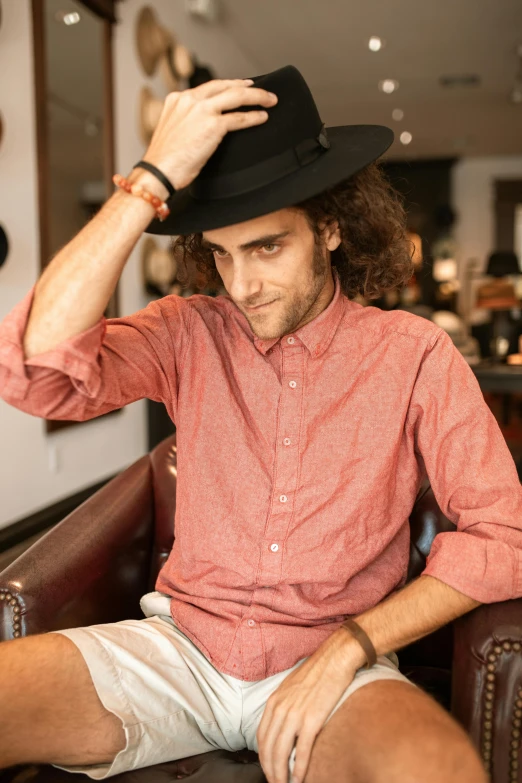 a man sitting in a chair with a hat on, long hair and red shirt, clear julian lage face, curly haired, wearing stripe shirt