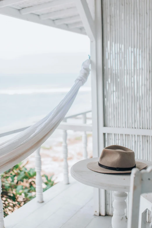 a hat sitting on top of a table next to a hammock, pexels contest winner, balcony door, serene beach setting, white, morning haze