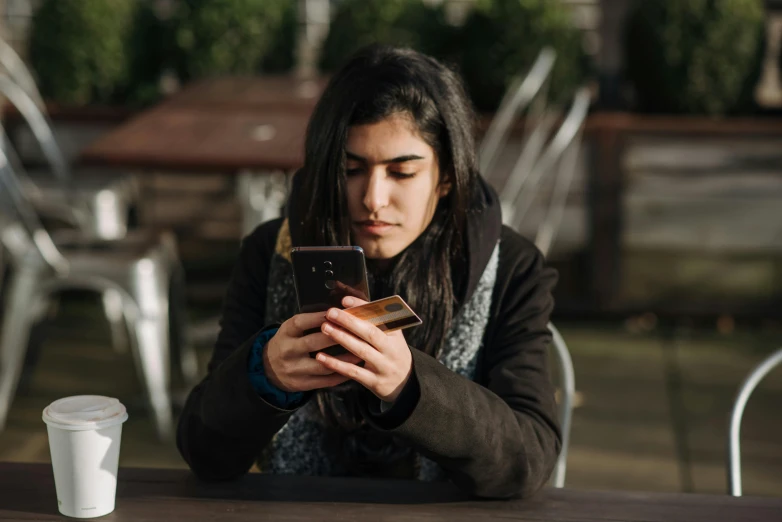 a woman sitting at a table looking at her cell phone, trending on pexels, happening, indian girl with brown skin, androgynous male, avatar image, outside