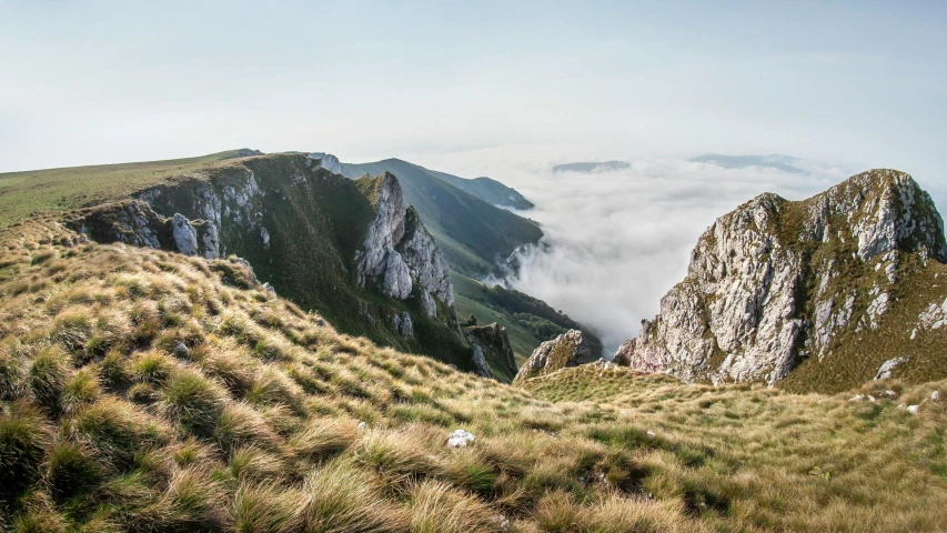 a man standing on top of a grass covered hillside, by Adam Marczyński, pexels contest winner, baroque, sheer cliffs surround the scene, covered in clouds, panoramic, fine art print