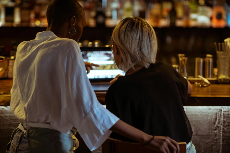 two women sitting at a bar looking at a laptop, by Lee Loughridge, trending on unsplash, happening, long shot from the back, standing elegantly, server in the middle, close up to the screen