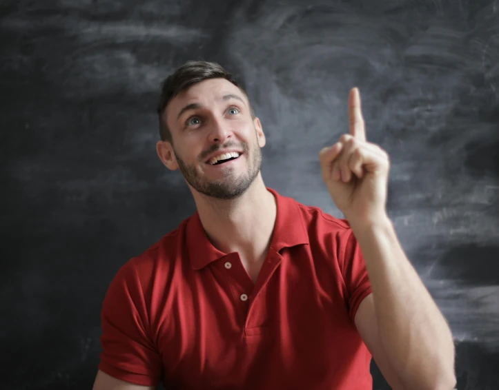 a man in a red shirt standing in front of a blackboard, pexels contest winner, pointing index finger, looking upwards, background image, smiling seductively