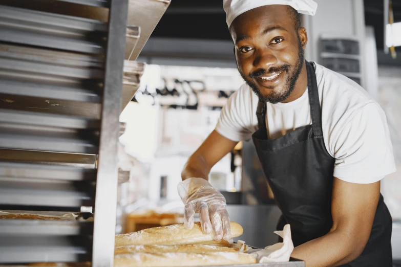 a man in an apron putting bread into an oven, pexels contest winner, african canadian, holding a baguette, people at work, a confident smile