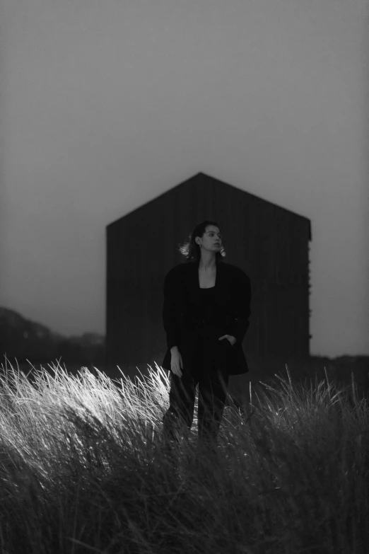 a woman standing in a field next to a building, a black and white photo, unsplash, conceptual art, black house, album cover, dimly - lit, coastal