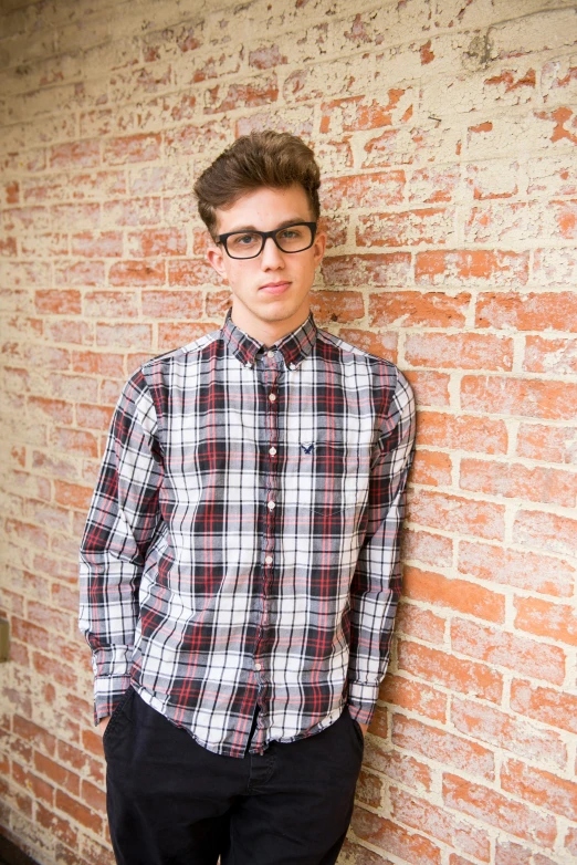 a young man leaning against a brick wall, an album cover, inspired by Harry Morley, plaid shirt, wearing eyeglasses, official store photo, square