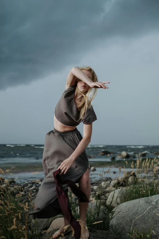 a woman standing on a rock near a body of water, inspired by Louisa Matthíasdóttir, unsplash, renaissance, brutal shapes stormy sky, doing a sassy pose, dressed with long fluent clothes, nordic summer