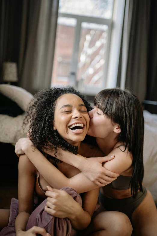 a couple of women sitting on top of a bed, pexels contest winner, incoherents, kissing smile, mixed-race woman, both laughing, hugging each other