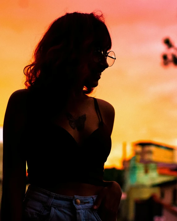 a woman with glasses standing in front of a sunset, inspired by Elsa Bleda, trending on pexels, wearing a cropped black tank top, bisexual lighting, trans rights, low angle photo