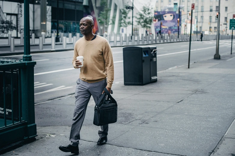 a man walking down a street holding a cup of coffee, lance reddick, grey pants and black dress shoes, holding a gold bag, wearing a black sweater