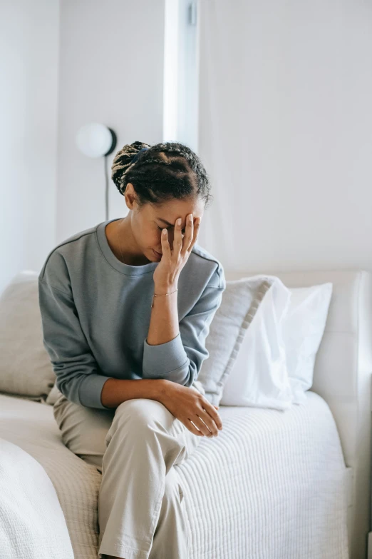 a woman sitting on a bed with her head in her hands, trending on pexels, with a hurt expression, sitting on couch, ashteroth, reassuring