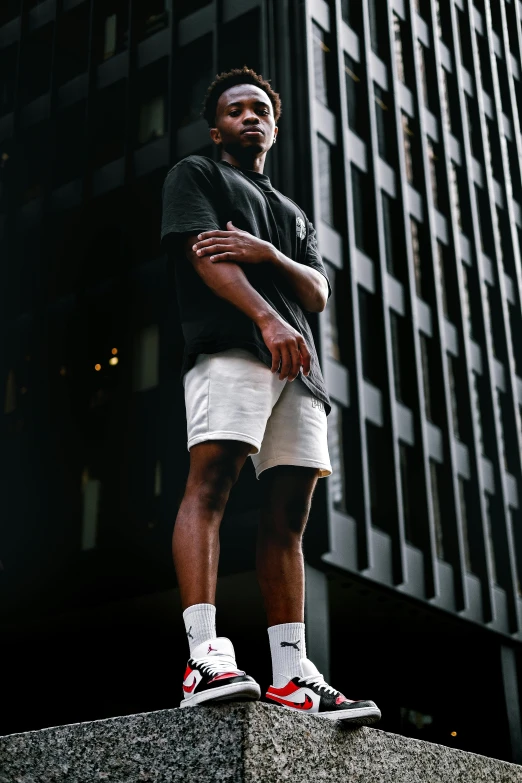 a man standing in front of a tall building, bra and shorts streetwear, dark-skinned, front lit, white and black clothing