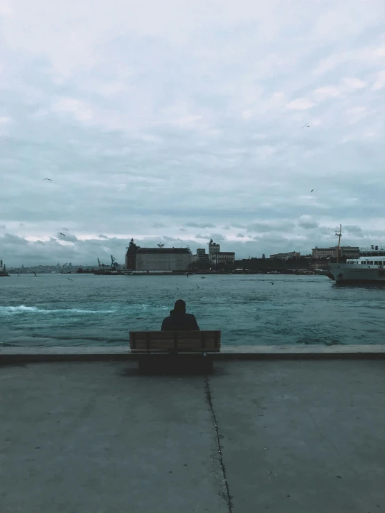 a person sitting on a bench in front of a body of water, inspired by Elsa Bleda, pexels contest winner, hyperrealism, harbour in background, overcast skies, trending on vsco, venice biennale