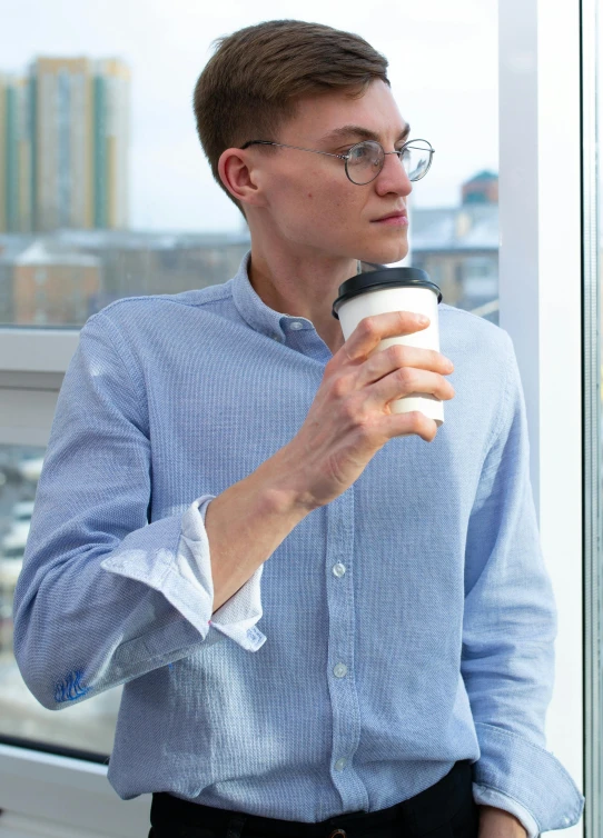 a man standing next to a window holding a cup of coffee, wearing a light blue shirt, non-binary, office clothes, pondering