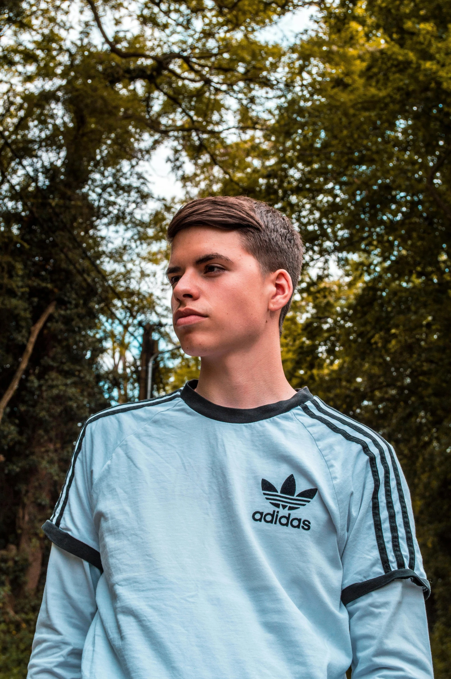 a young man standing in front of a forest, an album cover, by Jacob Toorenvliet, unsplash, realism, wearing adidas clothing, headshot profile picture, declan mckenna, low quality photo