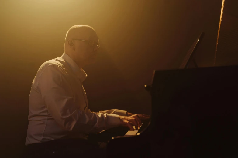 a man sitting at a piano in a dark room, bald man, man with glasses, during golden hour, performing a music video