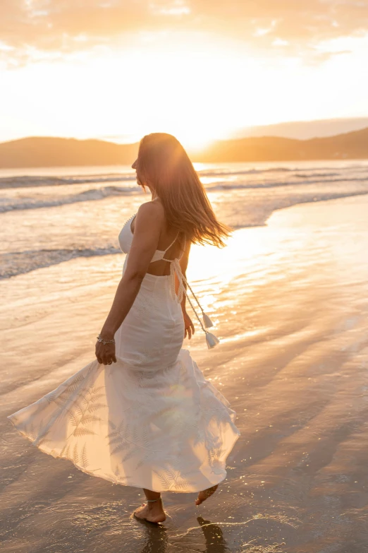 a woman in a white dress walking on a beach, sun down golden hour, abel tasman, flowing long hair, sun - drenched