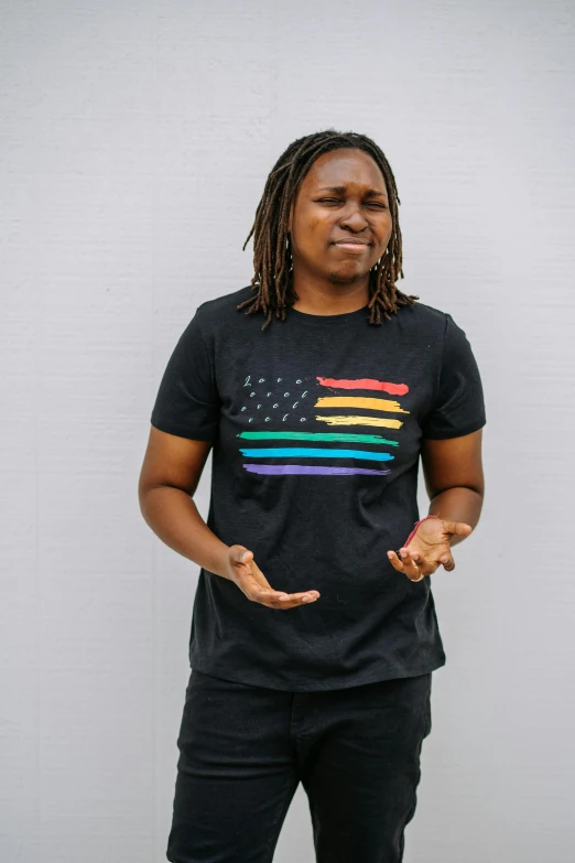 a man standing in front of a white wall, inspired by David Brewster, unsplash, black arts movement, pride flag in background, black t-shirt, 30 years old woman, stars and stripes