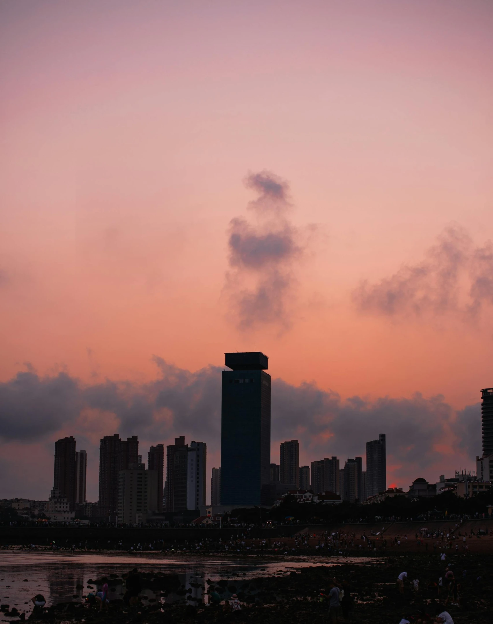 a group of people standing on top of a beach next to a body of water, an album cover, pexels contest winner, colombo sri lanka cityscape, pink skies, chimneys on buildings, humid evening