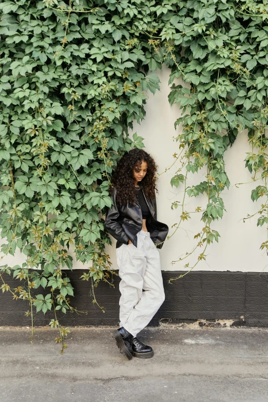 a woman leaning against a wall covered in vines, an album cover, inspired by Esaias Boursse, trending on unsplash, baggy jeans, tessa thompson inspired, leather jackets, wearing white sneakers