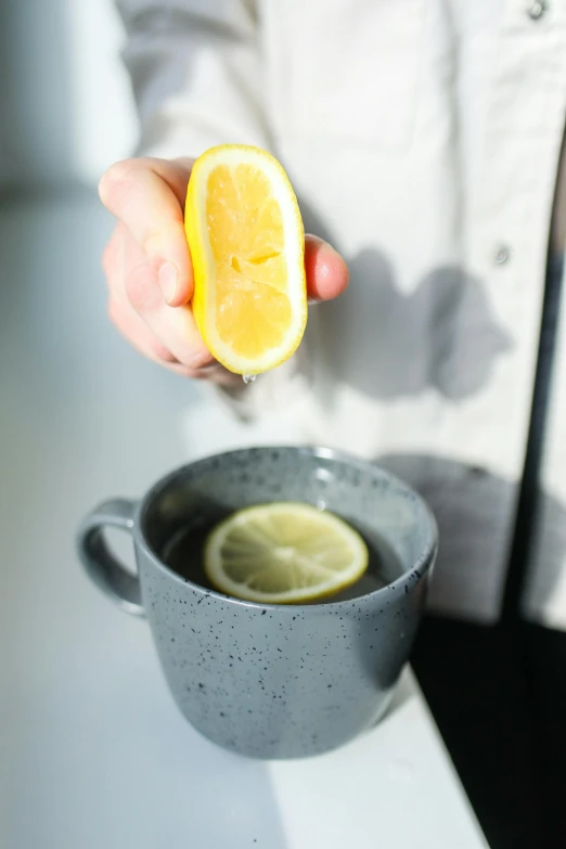 a person squeezing a lemon into a cup of tea, by Alice Mason, grey, indoor shot, displayed, hydration