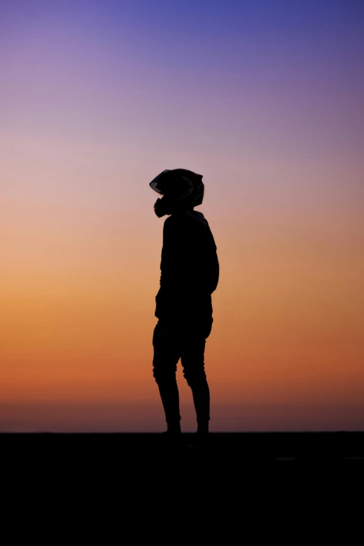 a man standing on top of a grass covered field, by Julian Allen, pexels, single silhouette figure, balaclava covering face, standing on the beach at sunset, avatar image