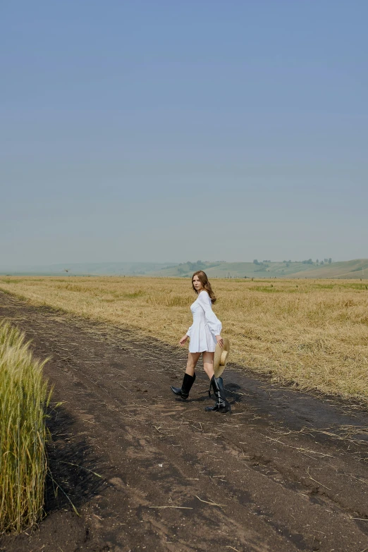 a woman in a white dress walking down a dirt road, in the middle of a field, knee-high boots, zezhou chen, color photograph