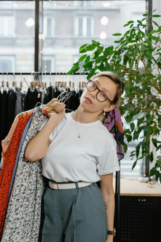 a woman standing in front of a rack of clothes, wearing round glasses, foliage clothing, very expressive, commercially ready