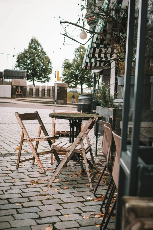 a couple of wooden chairs sitting on top of a brick sidewalk, by Jesper Knudsen, unsplash, happening, table in front with a cup, old shops, seasonal, in a square