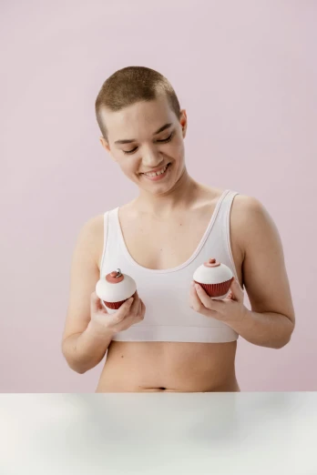 a woman in a white tank top holding two cupcakes, by Matija Jama, trending on pexels, renaissance, shaved head, with a large breasts, anatomically accurate, non binary model