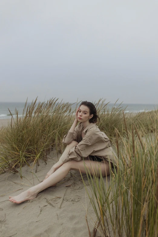 a woman sitting on top of a sandy beach, wearing an oversized sweater, standing in tall grass, pale skin, photoshoot for skincare brand