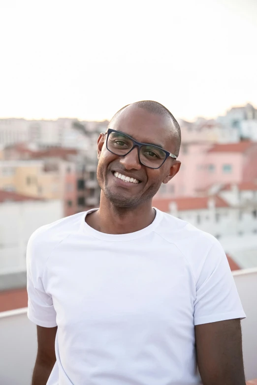 a man standing on top of a roof next to a building, inspired by Francis Souza, happening, happily smiling at the camera, dark skin tone, with glasses, lisbon