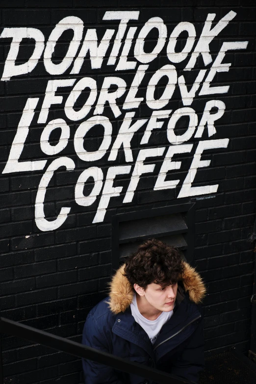 a man sitting in front of a sign that says don't look for one look for coffee, an album cover, inspired by John Luke, graffiti, declan mckenna, profile image, curls on top, shot from a drone
