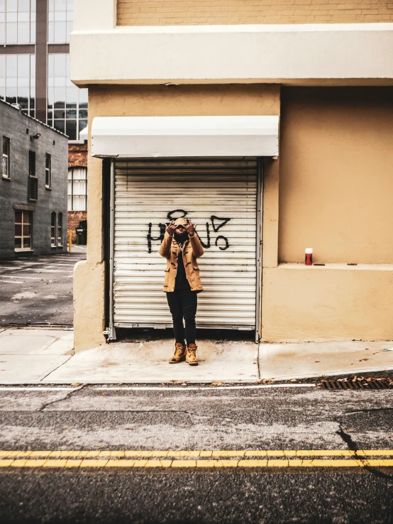 a man standing in front of a garage door, a photo, inspired by Elsa Bleda, trending on unsplash, graffiti, mysterious coffee shop girl, posing in disguise among humans, light brown coat, holding a camera