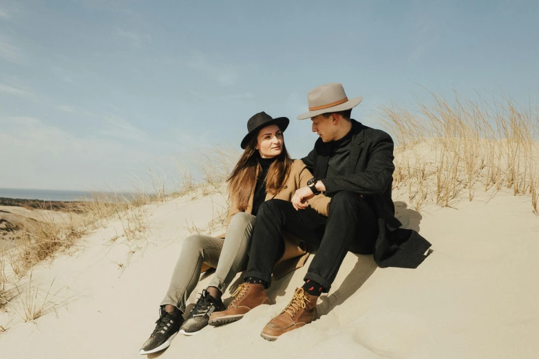 a man and a woman sitting on top of a sandy beach, by Emma Andijewska, trending on pexels, leather clothing and boots, beige fedora, avatar image, black