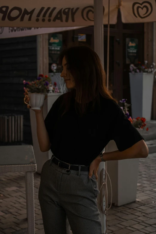 a woman standing under an umbrella on a sidewalk, wearing a black t-shirt, drinking a cup of coffee, standing in a dimly lit room, portrait featured on unsplash