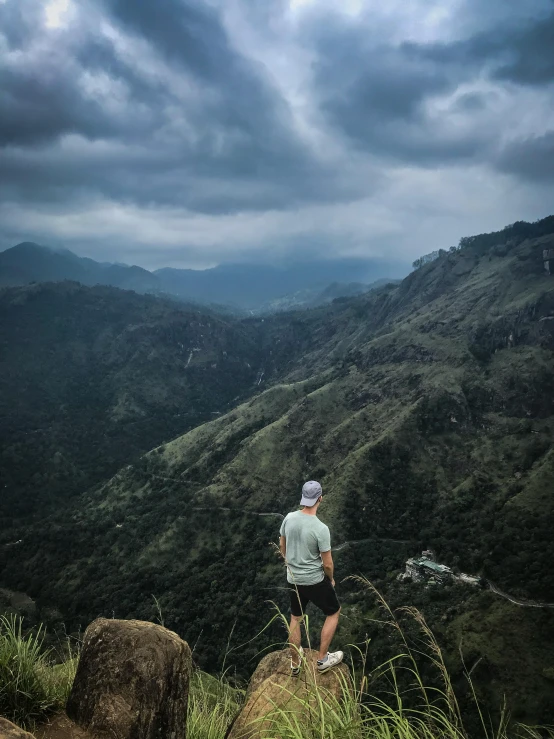 a man standing on top of a lush green hillside, a picture, happening, sri lankan landscape, dark and moody atmospheric, instagram post, with mountains in the distance