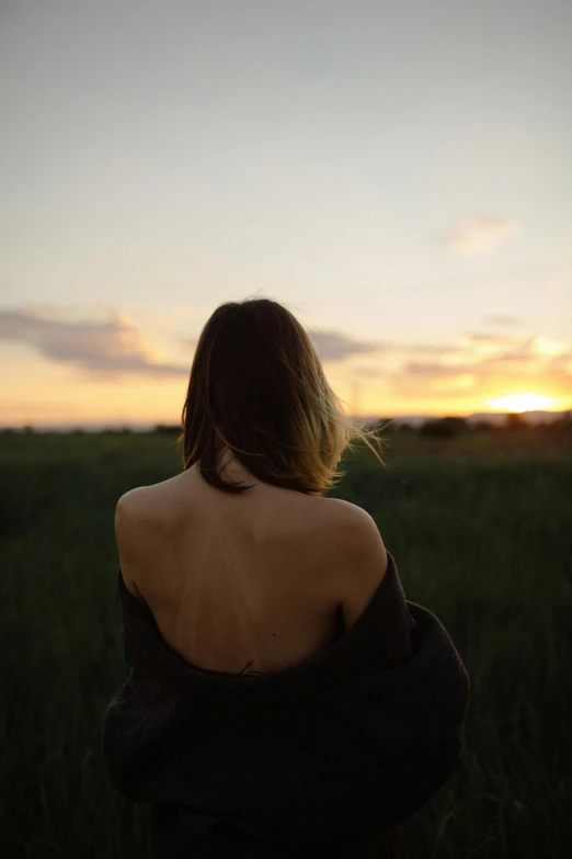 a woman sitting in a field at sunset, unsplash, showing her shoulder from back, bare shoulders, looking out, ((sunset))