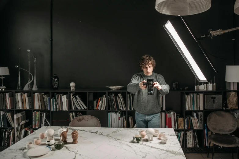 a man standing in front of a table holding a camera, by Jan Tengnagel, light and space, marble room, dark kitchen of an art student, ceramics, anton fadeev 8 k