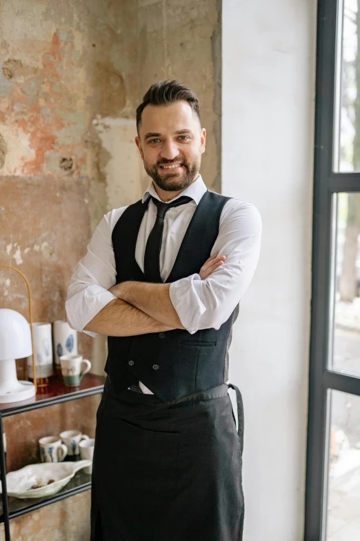 a man standing in front of a window with his arms crossed, a portrait, inspired by Ramon Pichot, pexels contest winner, bartending, wearing suit vest, happy chef, ukrainian