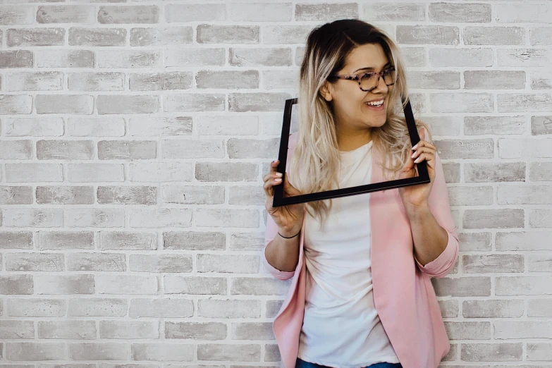 a woman standing in front of a brick wall holding a picture frame, inspired by Emma Ríos, pexels contest winner, square glasses, cute young woman, pokimane, a blond