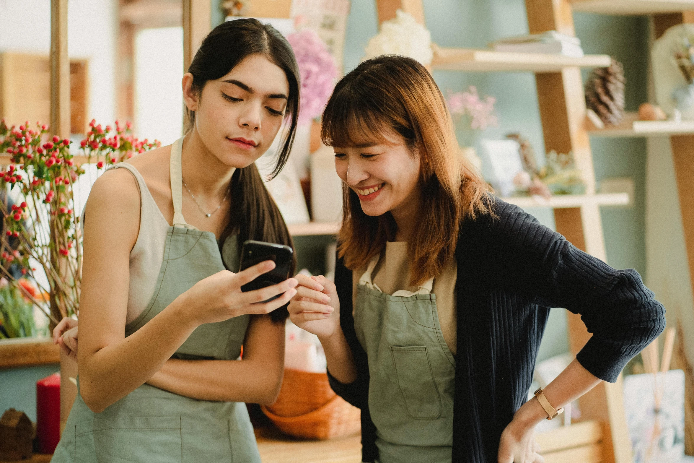 two women standing next to each other looking at a cell phone, trending on pexels, flower shop scene, avatar image