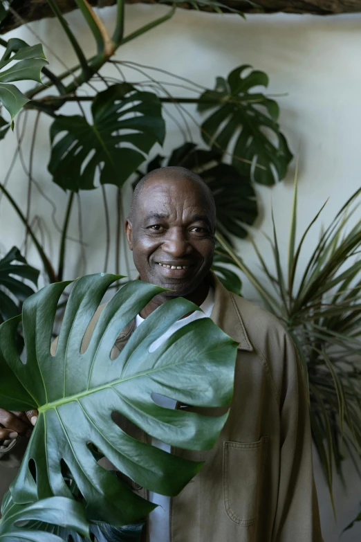 a man holding a large leaf of a plant, an album cover, inspired by Candido Bido, pexels contest winner, hurufiyya, while smiling for a photograph, huell babineaux, an elderly, big interior plants