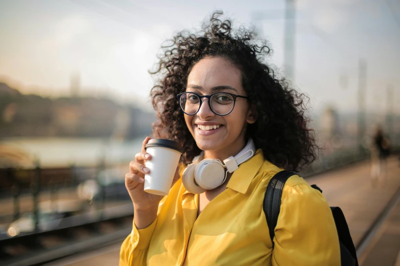 a woman standing on a train platform holding a cup of coffee, trending on pexels, wavy hair yellow theme, aida muluneh, with glasses on, student